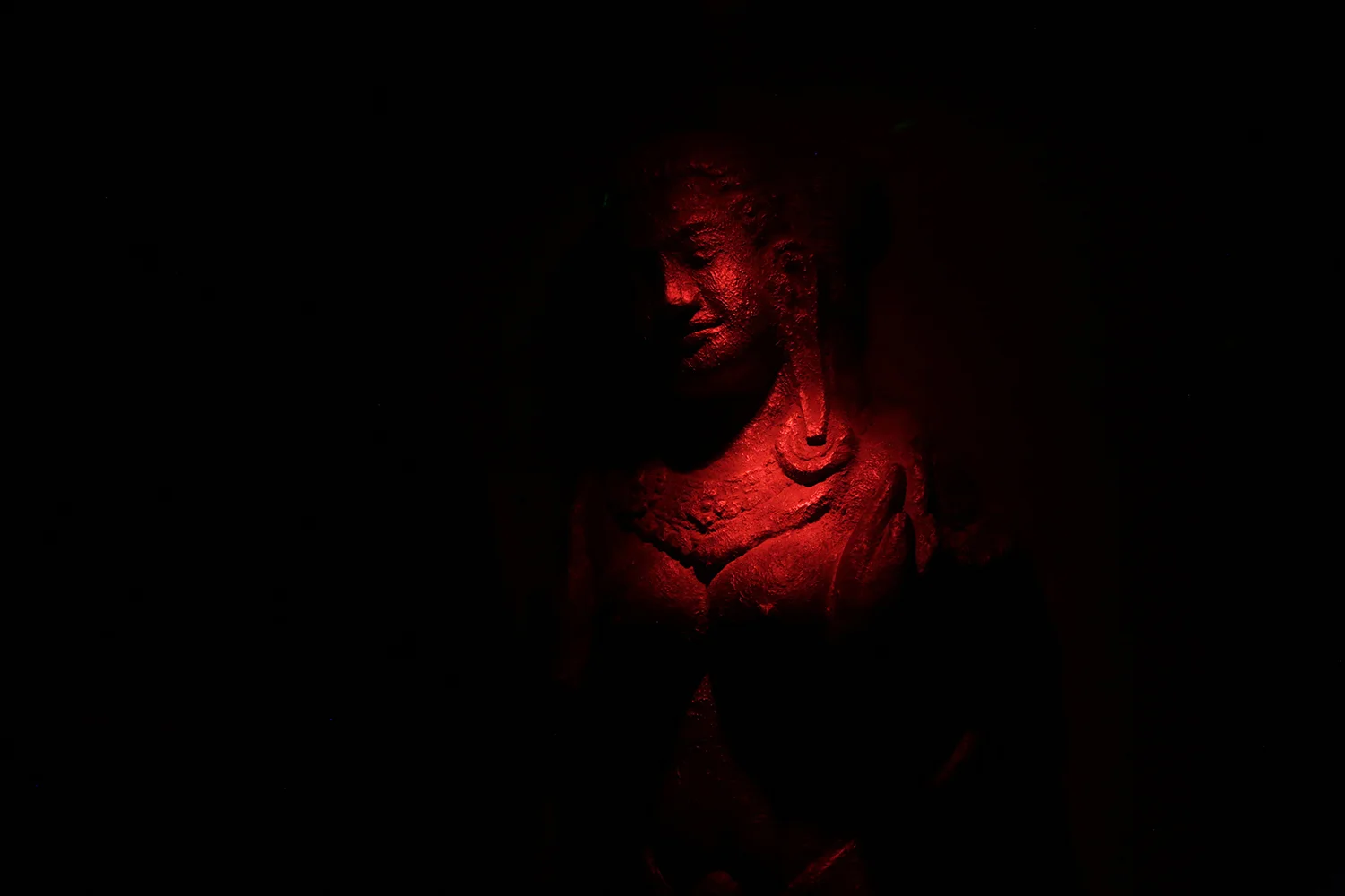 Analog photograph of a carved stone figure bathed in red light against total darkness, dramatic and mysterious mood with visible film grain