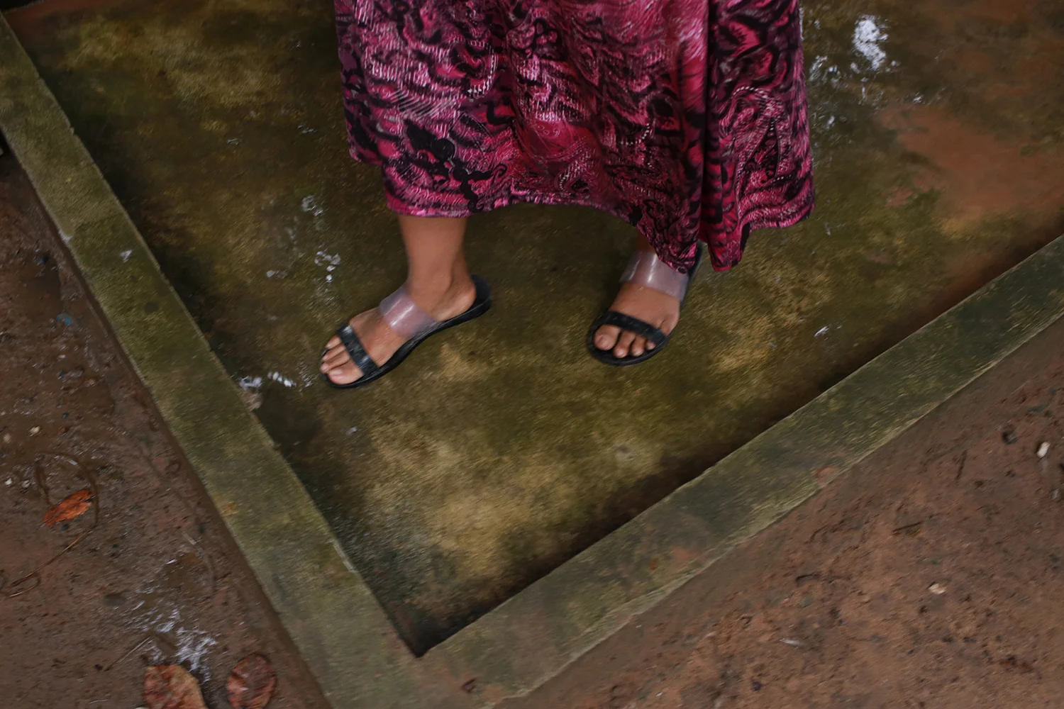 Analog close-up of a person's feet in black sandals and pink patterned skirt standing on wet mossy stone, earthy green and pink tones