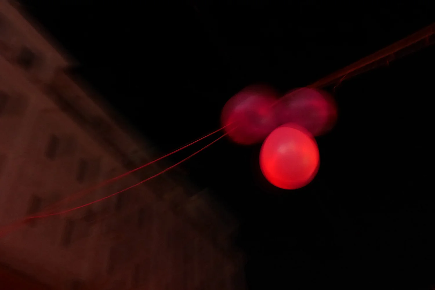 Moody analog photograph of three glowing red round lights against a dark night sky with a pale building edge, deep red and black tones